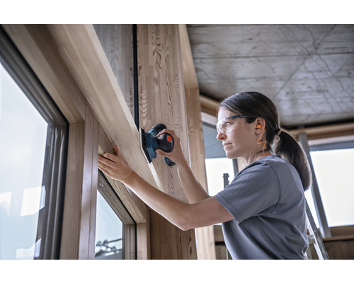 Frau schleift Holzfensterrahmen mit einem Schleifgerät, während sie Schutzbrille und Ohrstöpsel trägt.