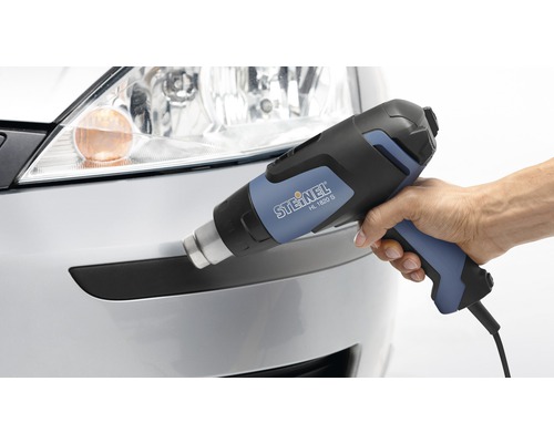 Main tenant un pistolet thermique STEINEL HL 1820 S devant un pare-chocs argenté