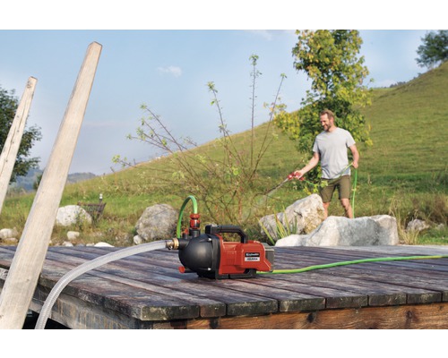 Einhell Hauswasserwerk auf einem Holzsteg mit einem Mann, der im Hintergrund mit einem Gartenschlauch gießt