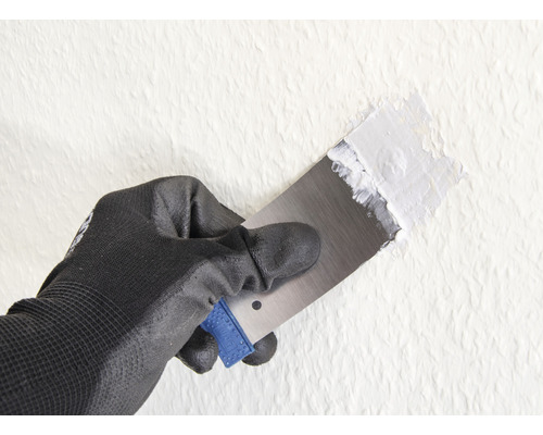 Hand mit Arbeitshandschuh und Spachtel trägt Spachtelmasse auf die Wand auf