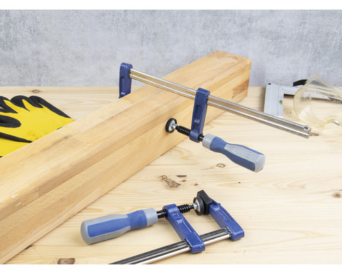 Zwei Schraubzwingen fixieren ein Holzbrett auf einem Holztisch, zusammen mit Arbeitshandschuhen, einem Tacker und einer Schutzbrille.