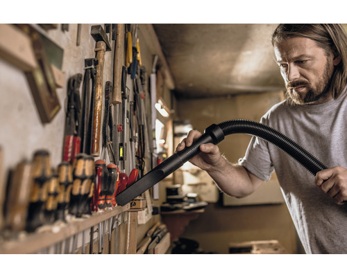 Handwerker reinigt Werkbank mit Staubsaugerdüse vor Werkzeugwand