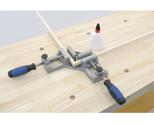 Eine Eckzwinge hält Holzleisten auf einer Werkbank zusammen, zusammen mit einer Flasche Kleber.