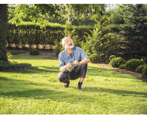 Un homme est accroupi sur une pelouse, un outil de jardinage à la main, et regarde vers le haut.
