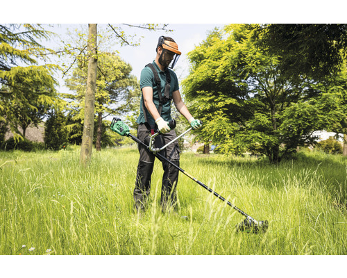 Mann mit Gesichtsschutz und Trimmer bei der Gartenarbeit im hohen Gras