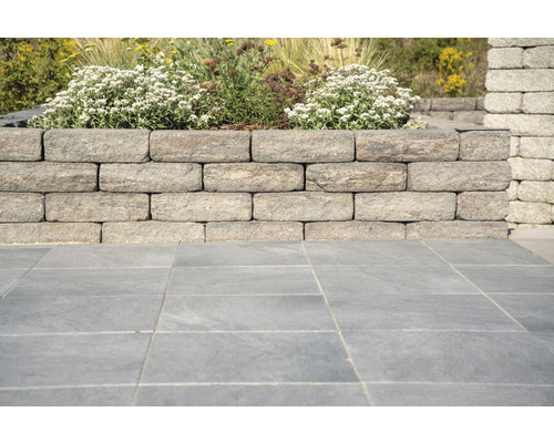 Eine Gartenmauer aus Steinblöcken mit Bepflanzung oben und Bodenplatten davor.