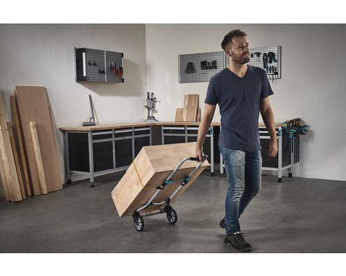 Mann transportiert Holz mit einem Transportwagen in einer Werkstatt mit Werkbank und Werkzeugwand