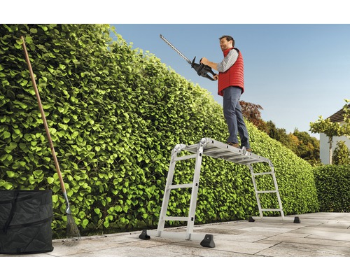 Un homme taille une haie avec un taille-haie depuis un échafaudage