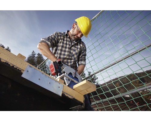 Un homme avec un casque de protection scie une planche de bois avec une scie circulaire Bosch sur un toit.