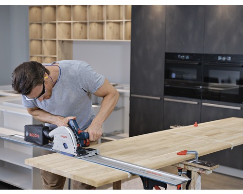 Un homme coupe une planche de bois avec une scie circulaire le long d'un rail de guidage dans un atelier de cuisine.
