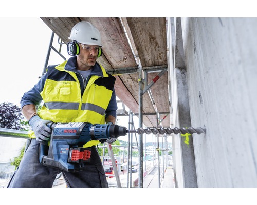 Un ouvrier du bâtiment perce un trou dans un mur en béton sur un échafaudage avec un marteau perforateur. Il porte des vêtements de protection et un casque.