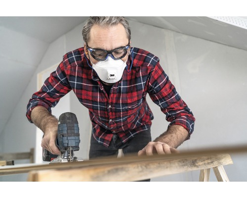 Homme travaillant avec une scie sauteuse, portant des lunettes de protection et un masque antipoussière.