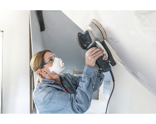 Une femme ponce un plafond avec une ponceuse excentrique, en portant un masque de protection et des lunettes de sécurité.