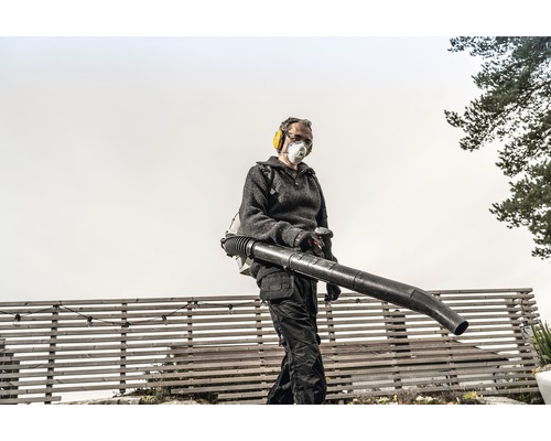 Une personne utilise un souffleur de feuilles dorsal dans le jardin, protégé par un casque antibruit et un masque anti-poussière.