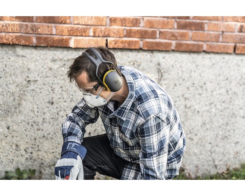 Un homme est agenouillé et porte un équipement de protection individuelle, notamment une protection auditive, des lunettes de sécurité, un masque respiratoire et des gants de travail.