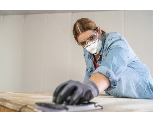 Une femme ponce du bois avec une cale à poncer, portant des lunettes de protection et un masque antipoussière.
