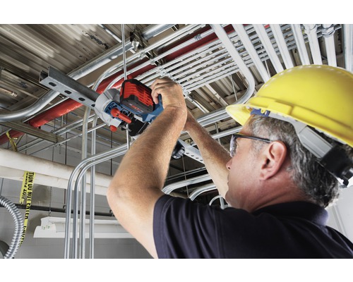 Un homme portant un casque de sécurité coupe un tuyau métallique au plafond avec une scie à tuyaux.