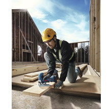 Ein Bauarbeiter mit Schutzhelm und Schutzbrille sägt ein Holzbrett mit einer Stichsäge auf einer Baustelle.