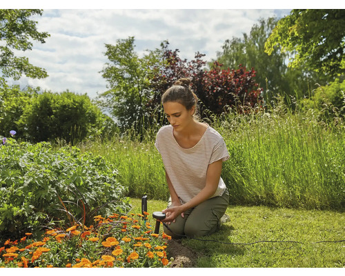 Eine Frau kniet im Garten und bedient einen Feuchtigkeitssensor zwischen Blumenbeeten