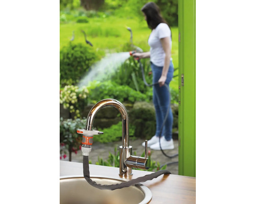 Gartenszene mit Wasserhahn und angeschlossenem Gartenschlauch, im Hintergrund eine Frau beim Bewässern von Pflanzen