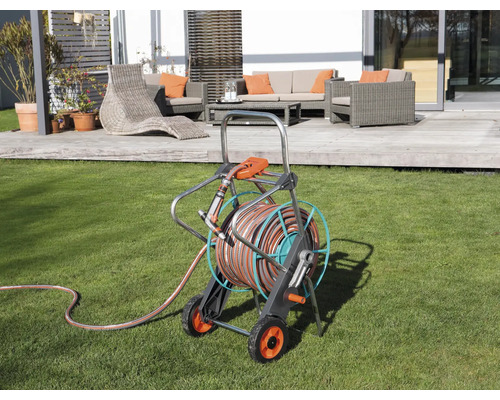 Gartenszene mit Schlauchwagen auf Rasenfläche und Terrassenmöbeln im Hintergrund.
