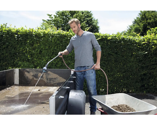 Un homme nettoie une remorque avec un tuyau d''arrosage et un pulvérisateur de jardin. Une brouette est posée à côté.