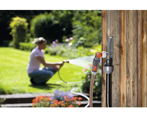 Gartenszene mit Schlauchverbinder und Wasserhahnanschluss an einer Holzwand, eine Frau bewässert den Garten