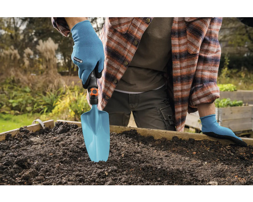 Personne portant des gants de jardinage utilisant une pelle de jardin dans un potager surélevé.