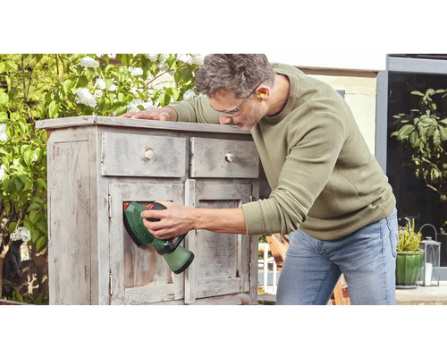 Mann schleift mit einem Schleifgerät ein Holzmöbelstück im Außenbereich.