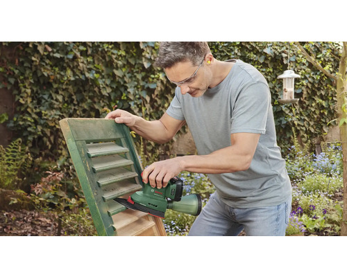 Ein Mann schleift mit einem Schleifgerät einen Fensterladen im Garten.