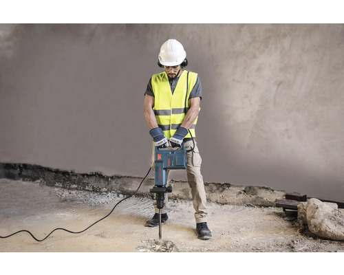 Un ouvrier du bâtiment portant un casque de protection, des lunettes de protection, une protection auditive, un gilet de sécurité et des gants utilise un marteau-piqueur.