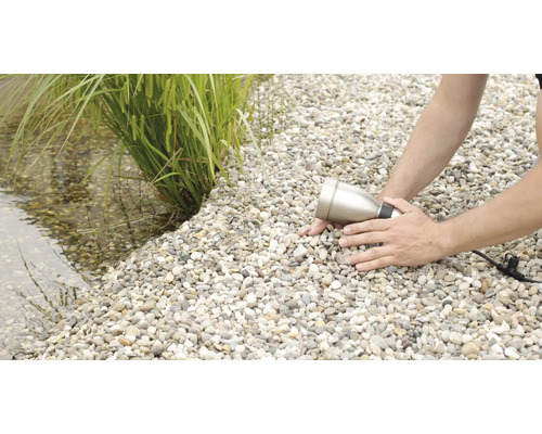 Ein Mann installiert einen Gartenstrahler auf einem Kiesweg neben einem Teich mit Pflanzen.