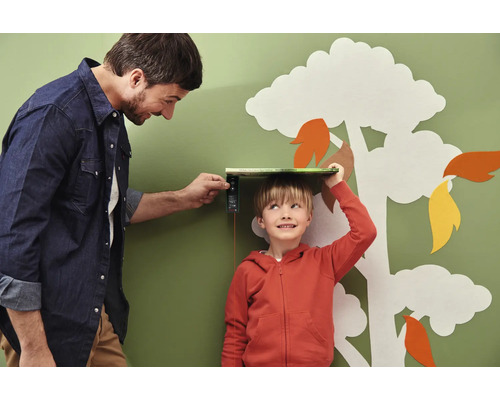 Un père mesure la taille de son fils avec un appareil de mesure numérique devant un mur avec une décoration d''arbre.