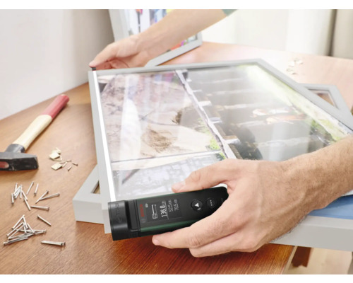 Un homme mesure un cadre photo avec un télémètre laser sur un établi avec un marteau et des clous.