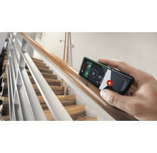 Une main tient un télémètre Bosch contre une rampe d''escalier en bois.