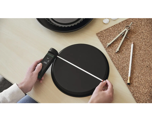 Une personne mesure le diamètre d''un moule à pâtisserie rond à l''aide d''un télémètre laser. Un deuxième moule à pâtisserie, un compas et un crayon sont posés sur la table.