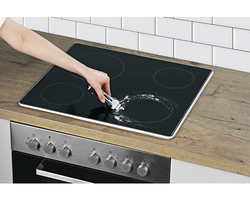 Eine Person reinigt ein Glaskeramikkochfeld mit einem Schaber in einer hellen Küche.