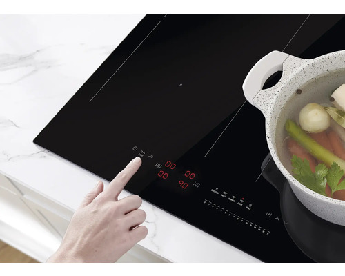 Eine Person bedient ein schwarzes Induktionskochfeld, auf dem ein Topf mit Gemüse steht.