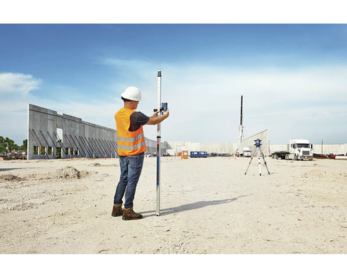 Ein Mann mit Bauhelm und Warnweste bedient ein Nivelliergerät auf einer Baustelle.