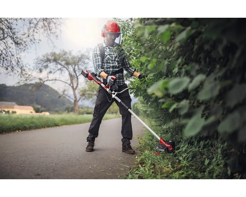 Une personne taille une haie avec un coupe-bordure sans fil porté avec une bandoulière. La personne porte des vêtements de protection.