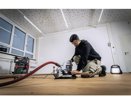 Ein Mann arbeitet mit einer Metabo Kapp- und Gehrungssäge in einem hellen Raum. Im Hintergrund sind ein Staubsauger und eine Baustellenleuchte zu sehen.