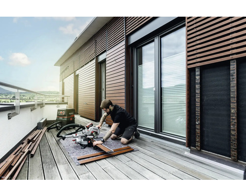 Mann arbeitet mit einer Kappsäge auf einem Balkon, um Holz zu bearbeiten. Ein Staubsauger steht bereit.