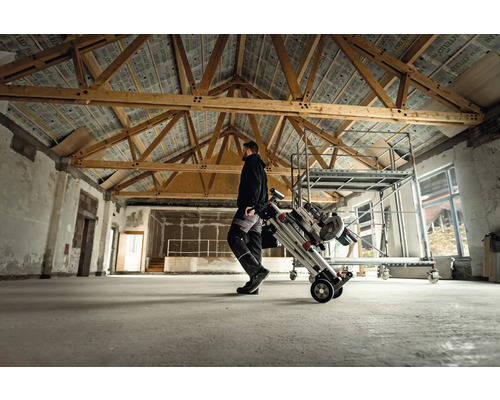 Un homme transporte un échafaudage roulant plié dans une pièce avec un plafond à poutres en bois.