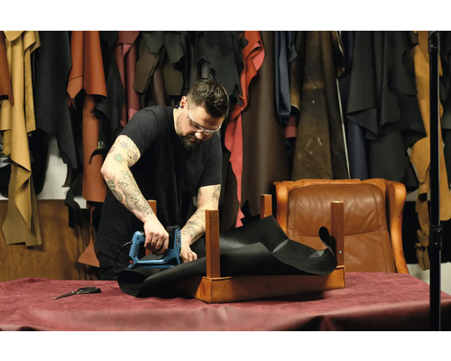 Ein Handwerker befestigt mit einem Elektrotacker ein schwarzes Stoffstück an einem Holzgestell in einer Werkstatt.