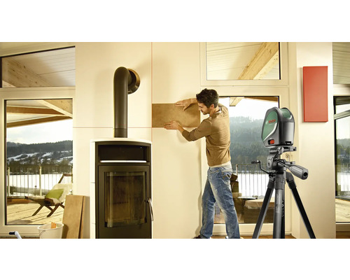 Mann verlegt Fliesen mit einem Kreuzlinienlaser zur Ausrichtung im Innenbereich neben einem Kamin.