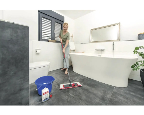 Salle de bains lumineuse avec baignoire îlot, toilettes et femme nettoyant le sol avec un balai à franges et un seau.