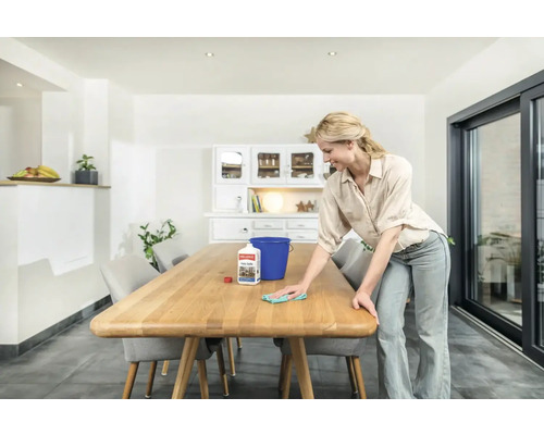 Une femme nettoie une table en bois avec un nettoyant et un chiffon dans une salle à manger lumineuse.