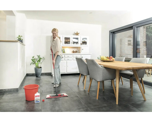 Femme nettoie le sol carrelé avec une serpillère, un seau et un nettoyant dans une salle à manger lumineuse