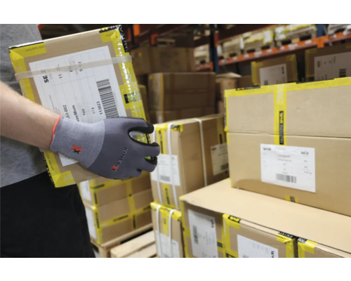 Eine Person mit Arbeitshandschuhen trägt einen Karton in einem Lager.