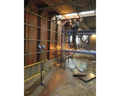 Un ouvrier debout sur un échafaudage coupe du métal avec une machine à découper, des étincelles jaillissant.